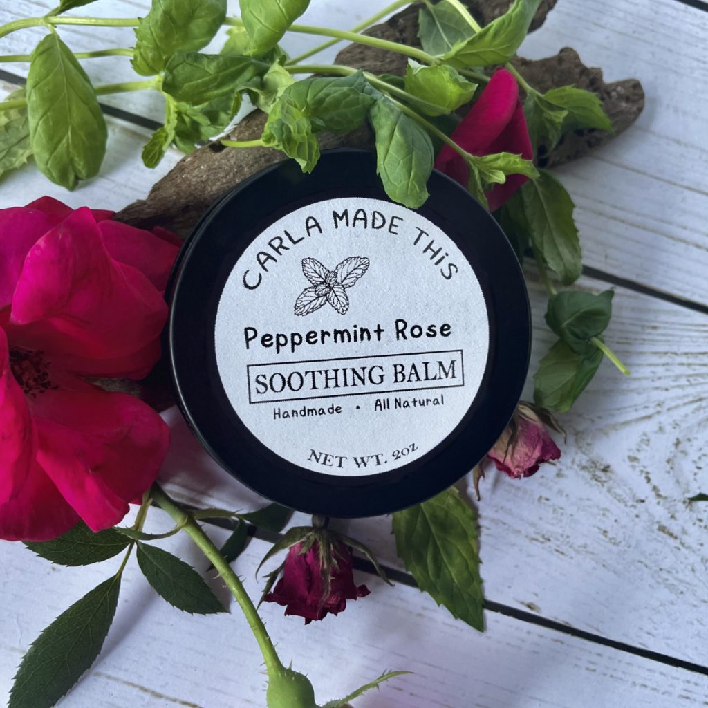 A black tin labelled peppermint rose soothing balm on a white wooden table in front of roses and bark.