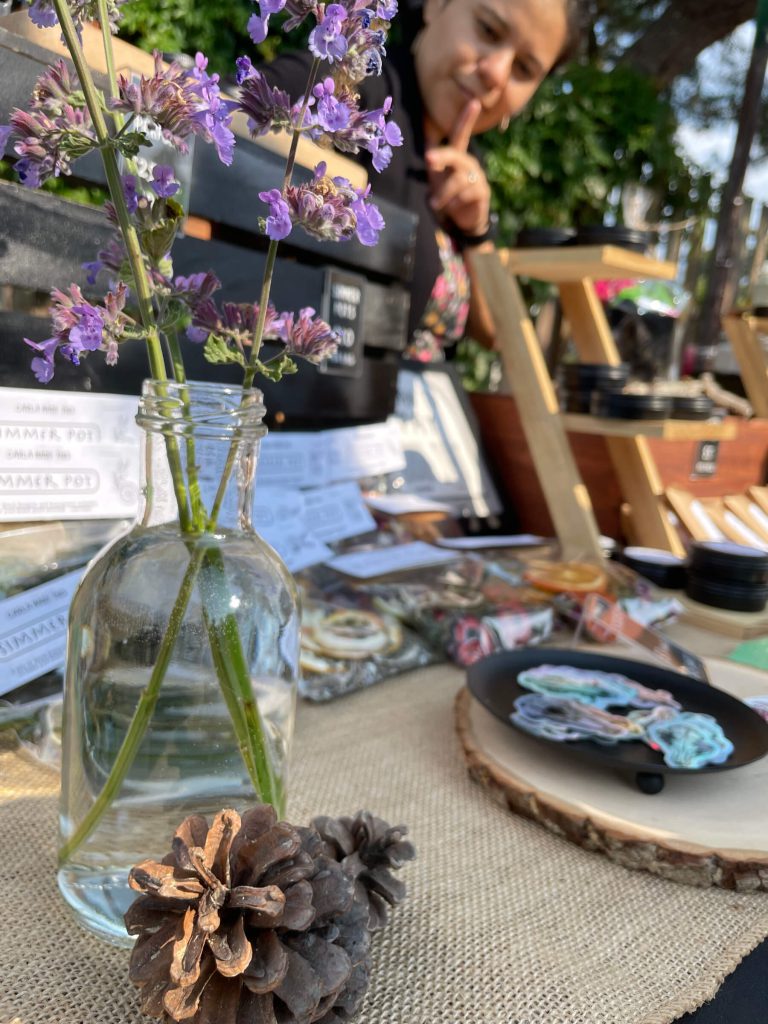 Some dried flowers in a clear vase sitting on the table in front of various products at a popup sale with Carla in the background.