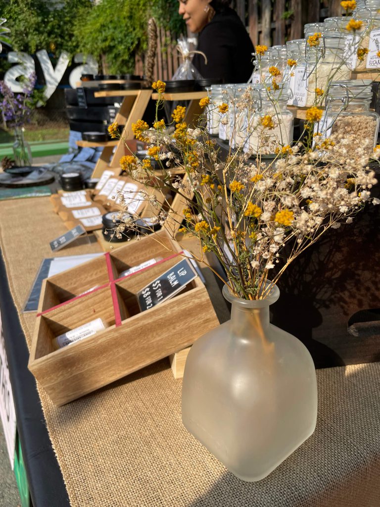 A bunch of dried flowers in a frosted vase sitting on the table in front of various products at a popup sale.