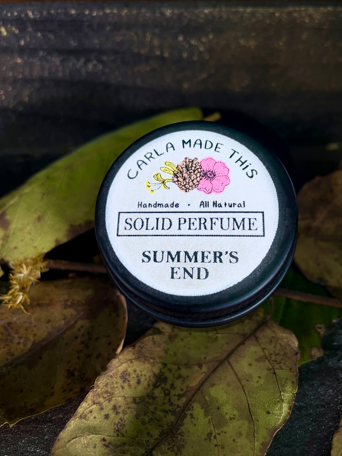 A small black container of summer's end solid perfume on a wooden table with leaves.