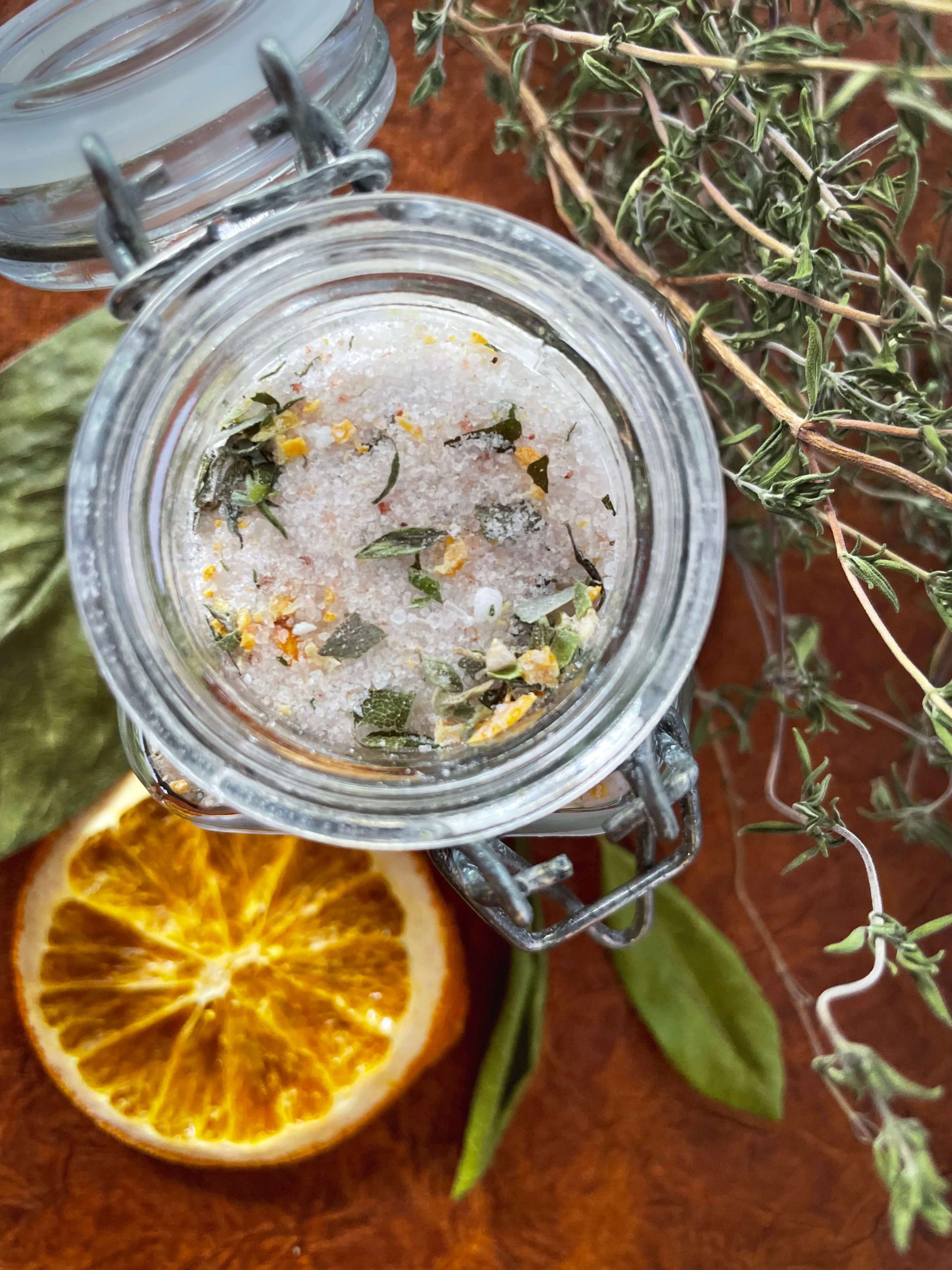 An open jar of wild bay orange thyme sitting on a table with herbs, citrus, and other items scattered below.