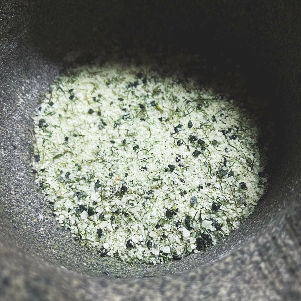 A closeup of a dark grey stone bowl containing white sea salt mixed with some black lava salt and fennel fronds.