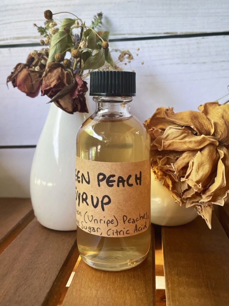 Bottle of green peach syrup in front of a vase of dried flowers