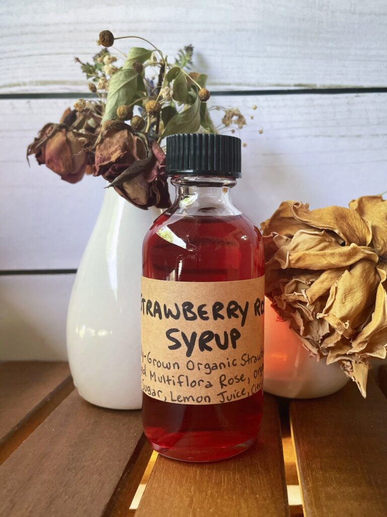 Bottle of strawberry rose syrup in front of a vase of dried roses.